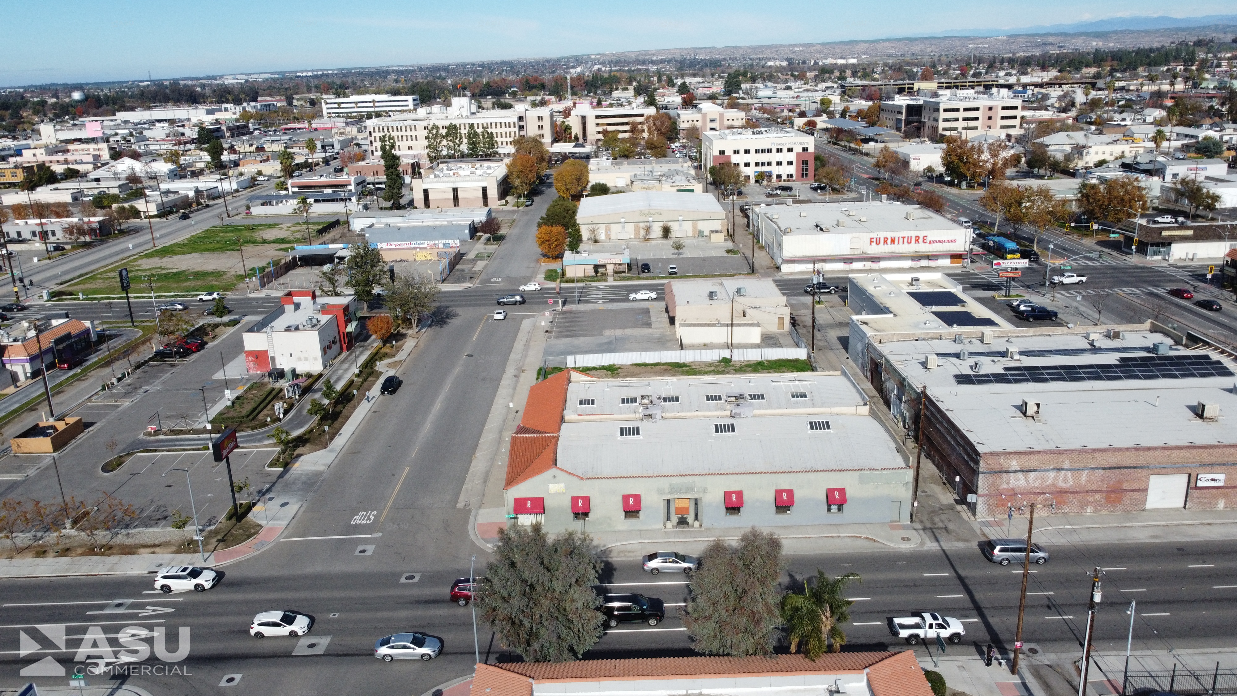 Oblique aerial of low commercial buildings, parking lots and vacant lots along a multi-lane street near mid-rise offices