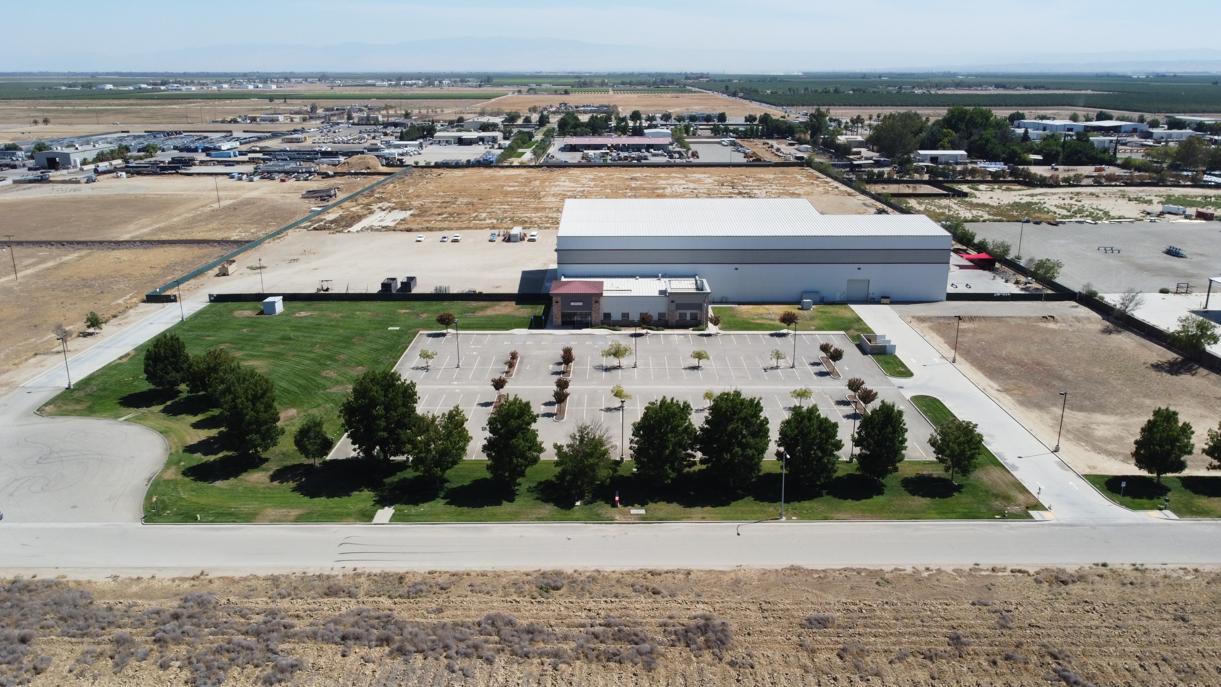 Aerial of an industrial building with a small office front, tree-lined parking lot, lawn and adjacent vacant land