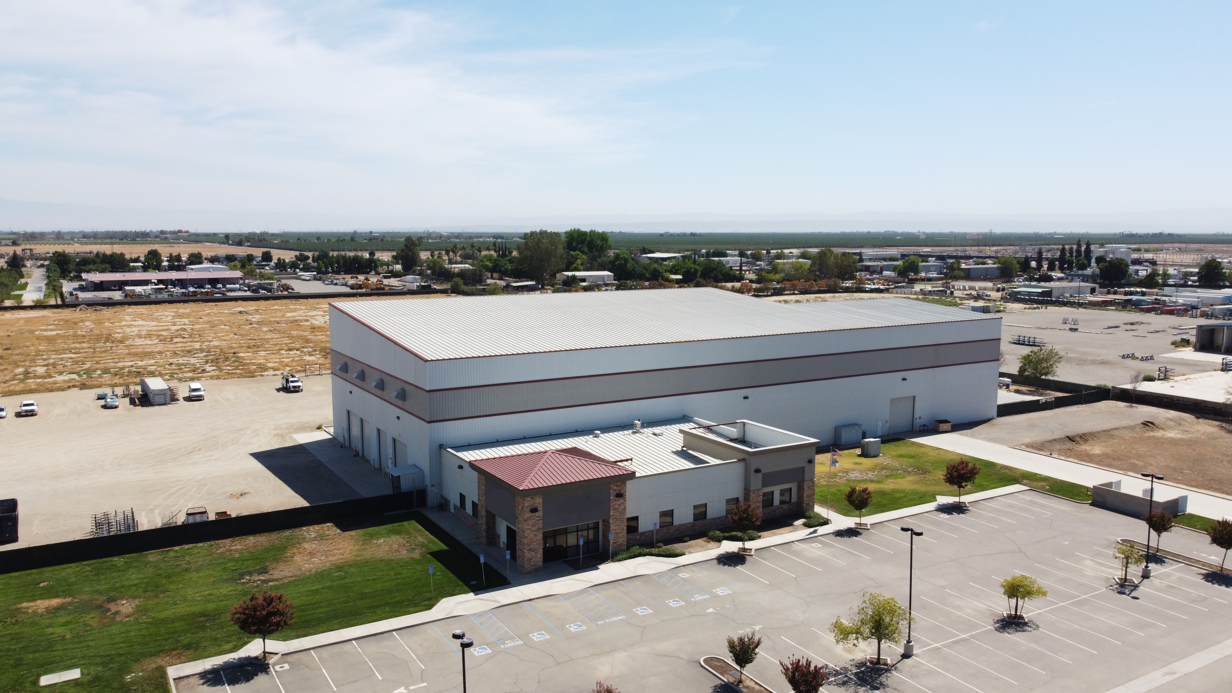 Aerial view of a large metal industrial building with office wing, parking lot and surrounding yards and orchards