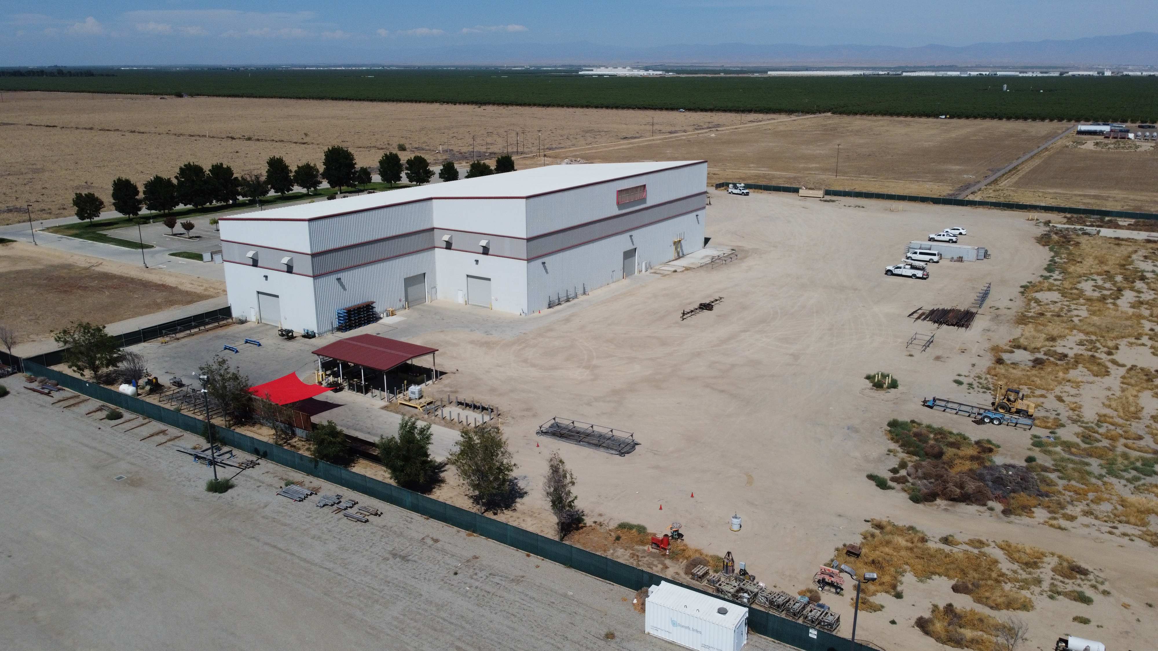 Aerial view of a large fenced industrial yard with a metal building, pipe racks, trailers and work trucks