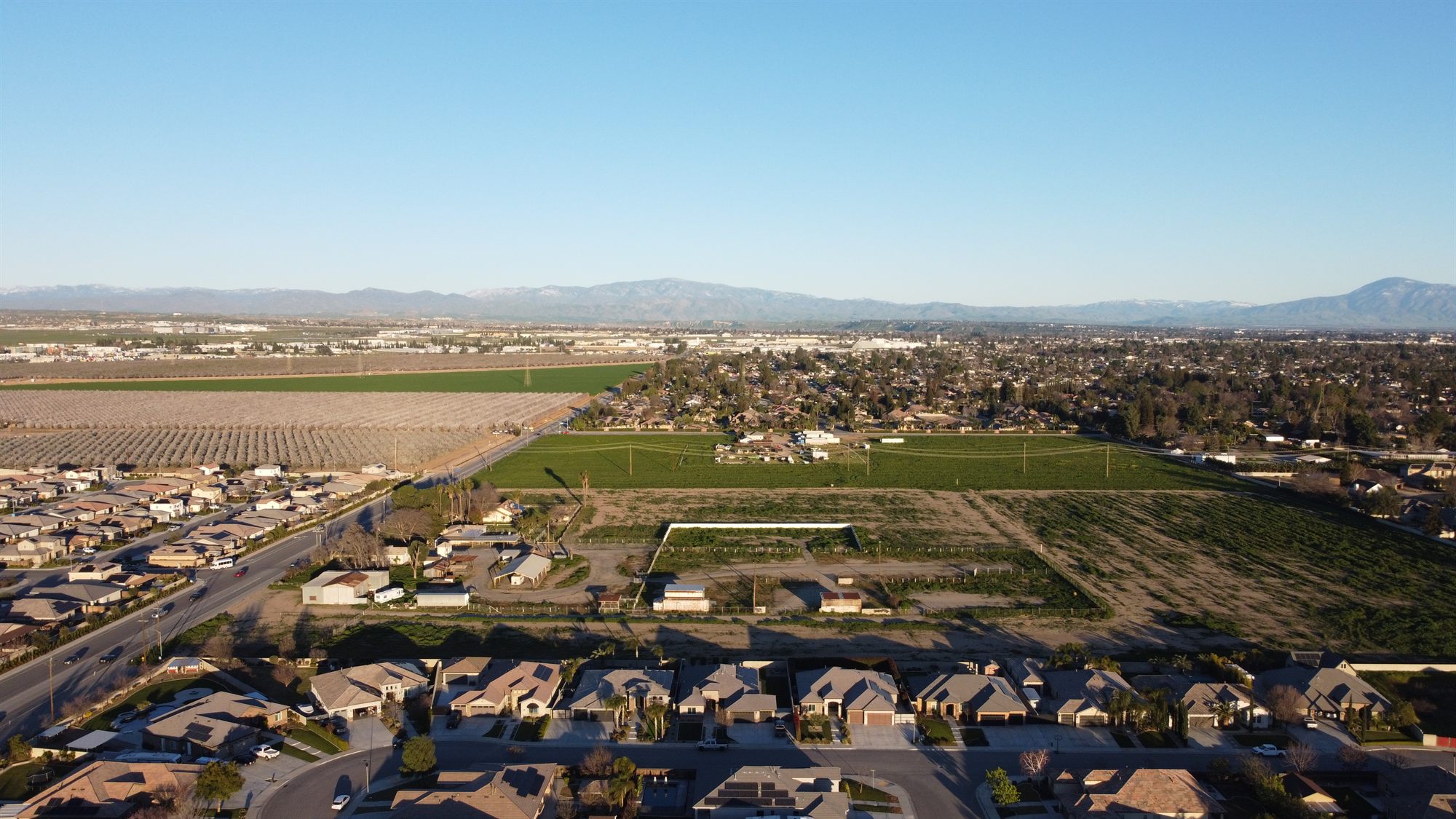 Aerial view of undeveloped land with small farm buildings beside new homes, orchards and crop fields