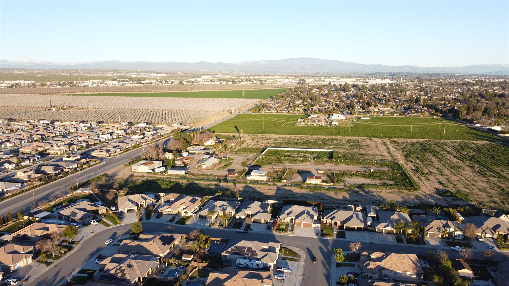 Aerial view of undeveloped land with a fenced parcel next to homes, a road, orchards and crop fields