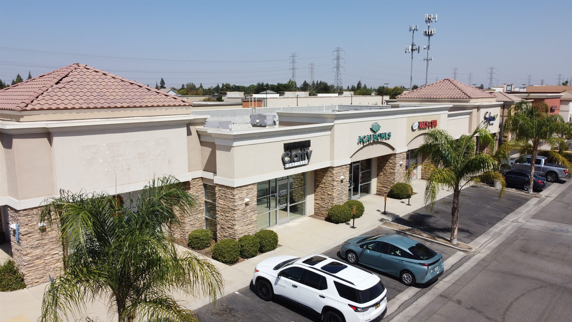 Elevated view of a one-story stucco and stone retail strip with tile-roof towers, palm trees and parked cars