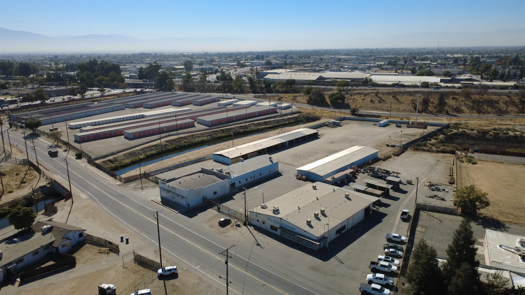 Aerial view of an industrial site with metal buildings and open sheds beside a canal and self-storage buildings