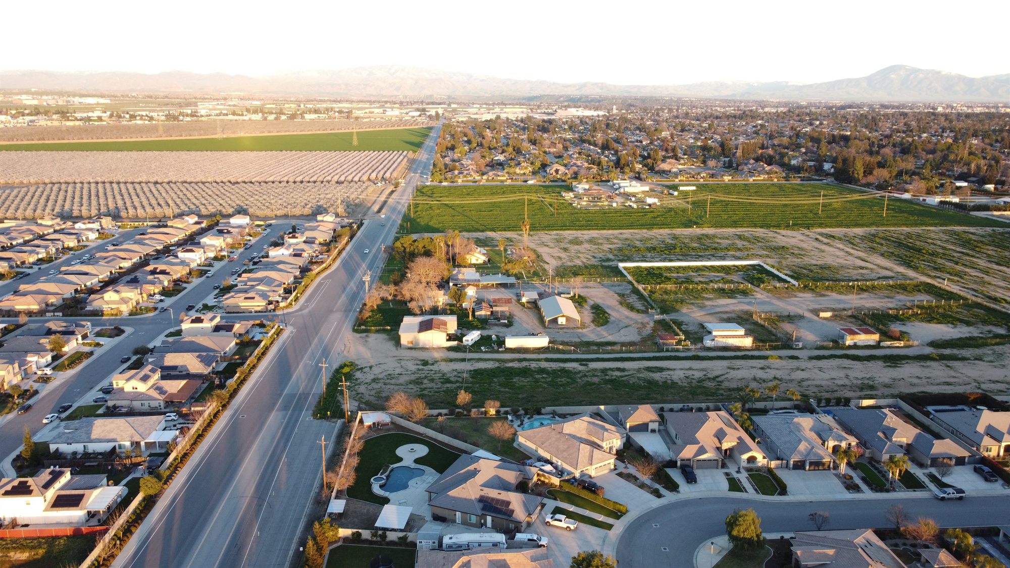 Aerial view of a large undeveloped parcel with farm buildings beside a road, homes, orchards and fields