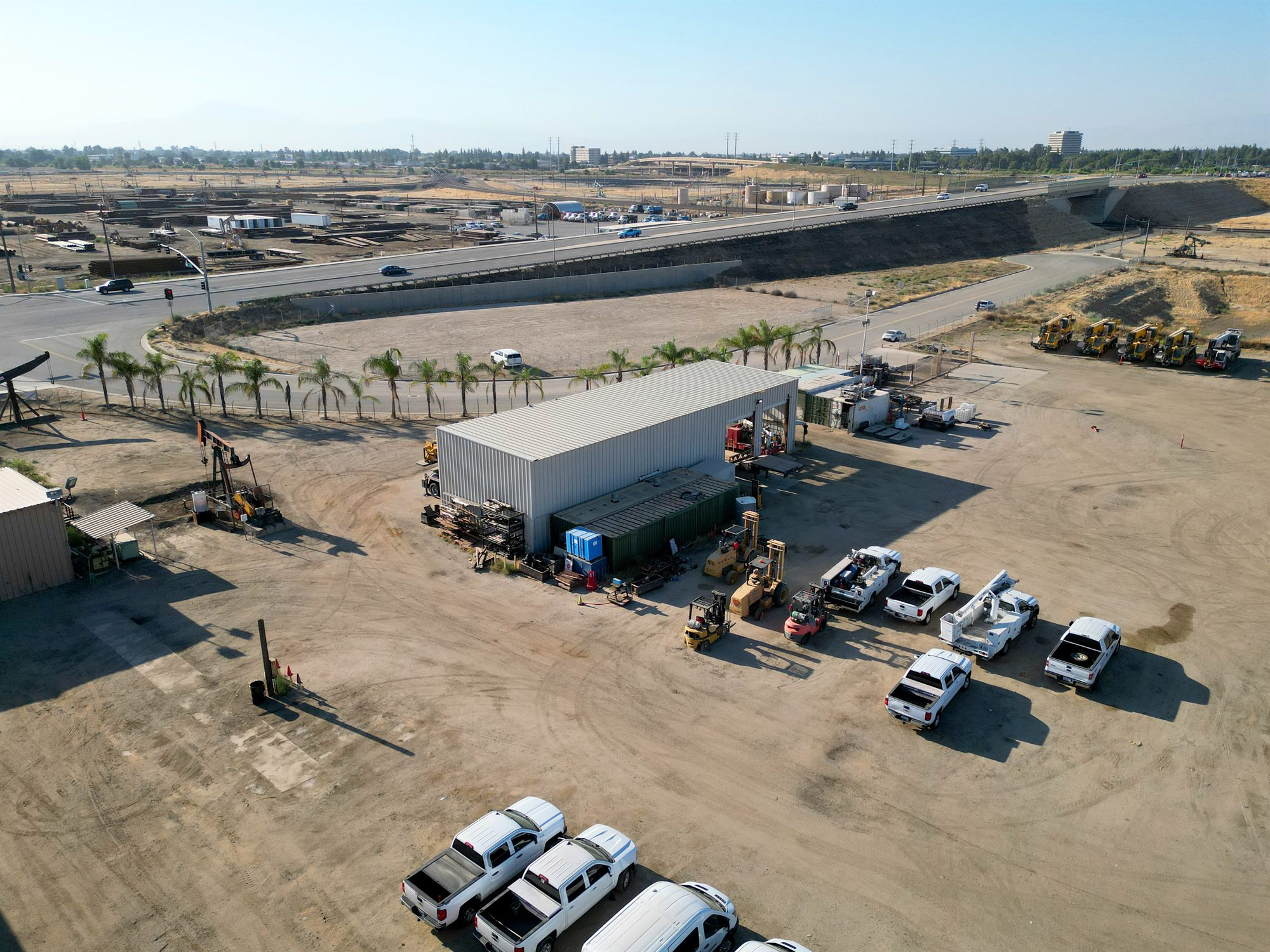 Oblique aerial of a metal shop building on a dirt yard with forklifts, service trucks and a pumpjack