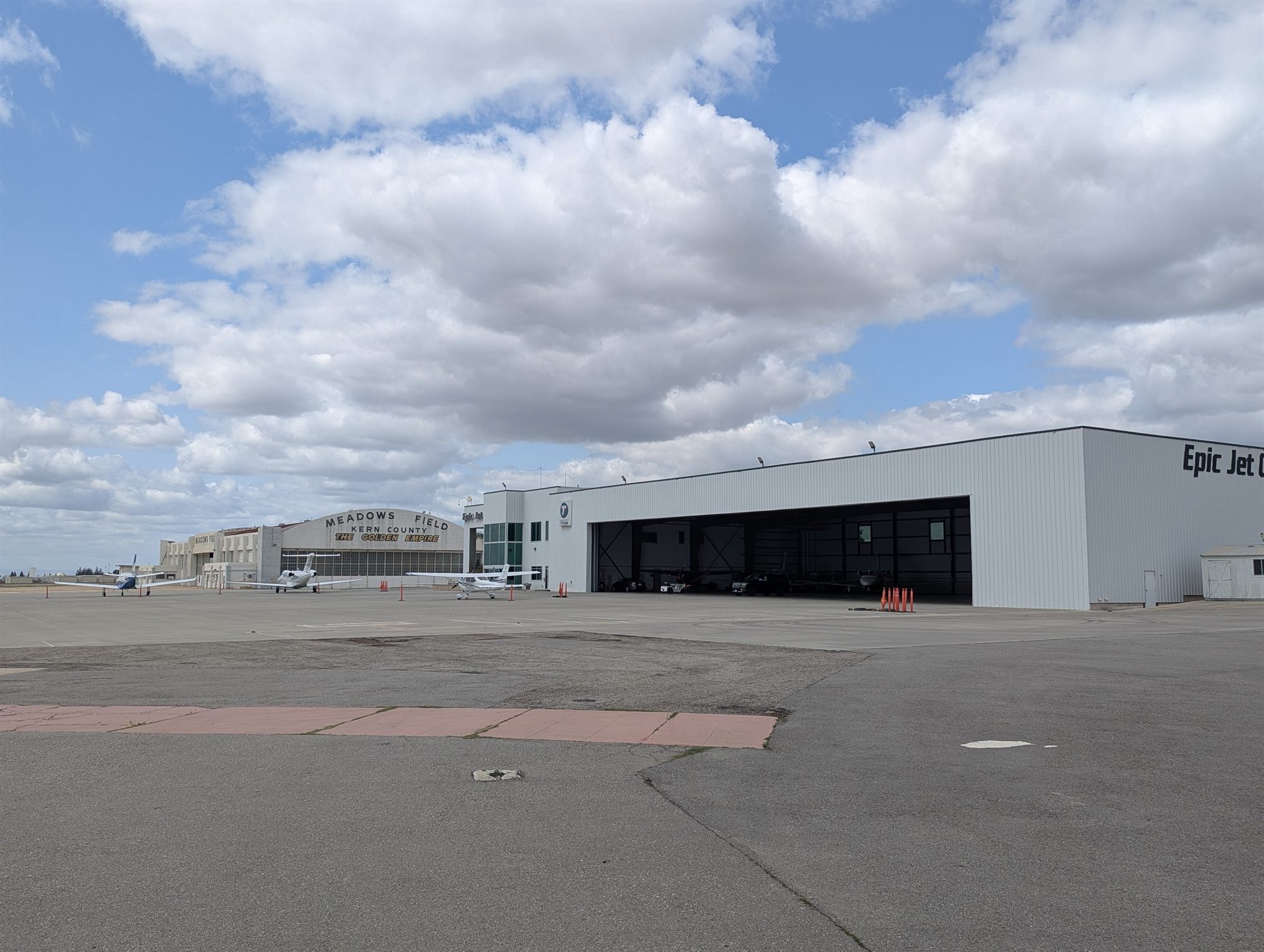 Airport apron with a large white metal hangar, a historic arched hangar behind and several small aircraft parked